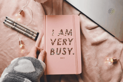 Ein Notizbuch mit Aufschrift I am very busy liegt auf einem Bett mit Laptop und Stiften.